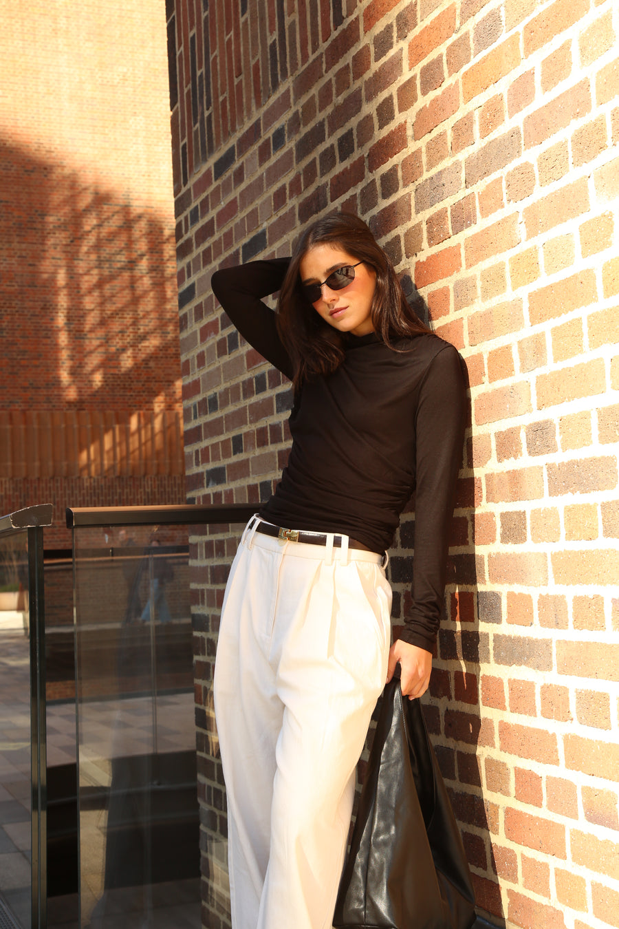 A model in a black long-sleeve top and cream wide-leg trousers, leaning against a sunlit brick wall
